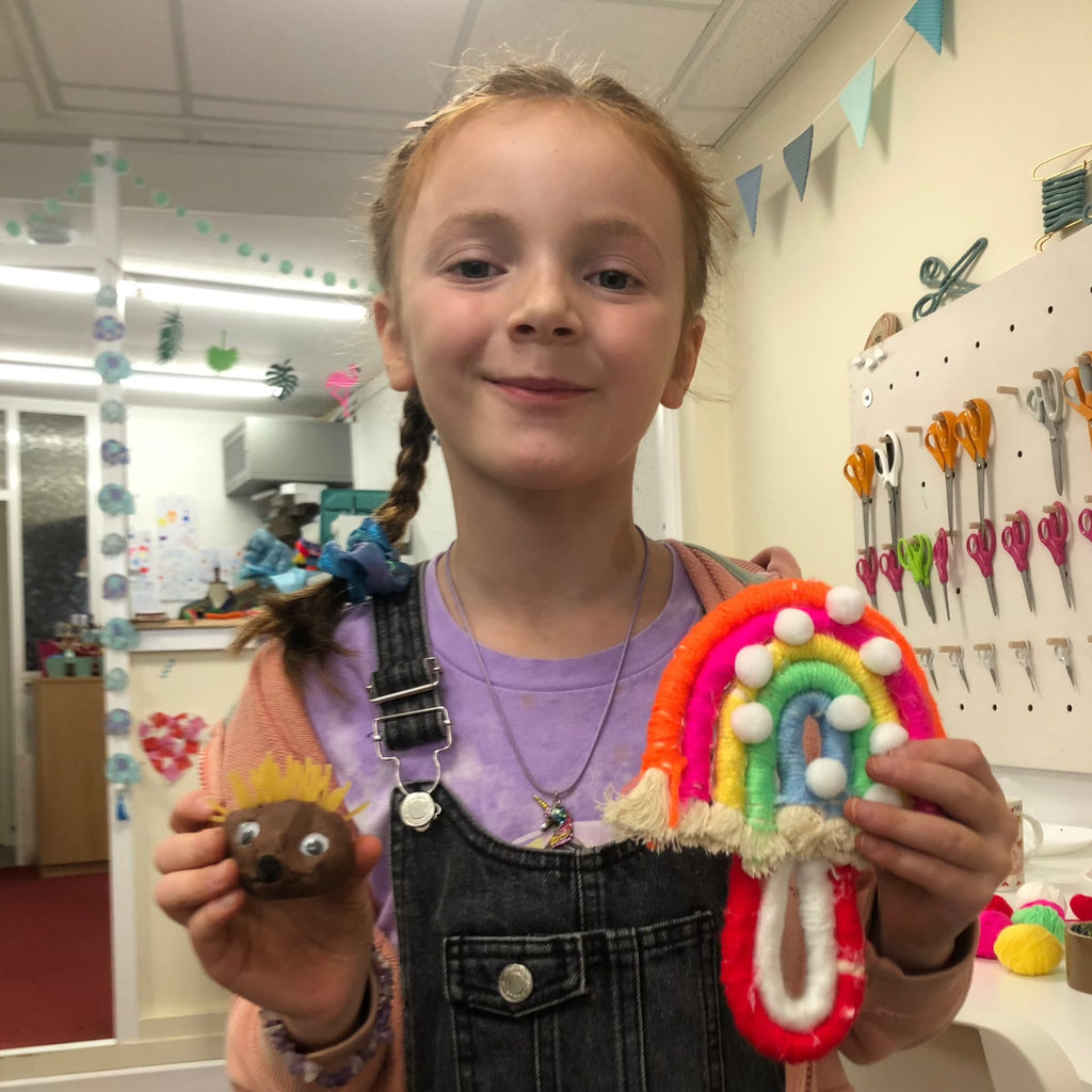 little girl with plaits holding her clay hedgehog and macrame rainbow she made at craft club.