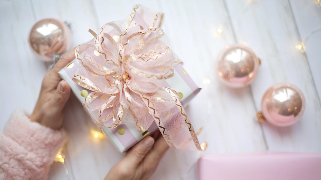beautifully packaged gift with pink bow, pink baubles and hands with white wooden floor background.
