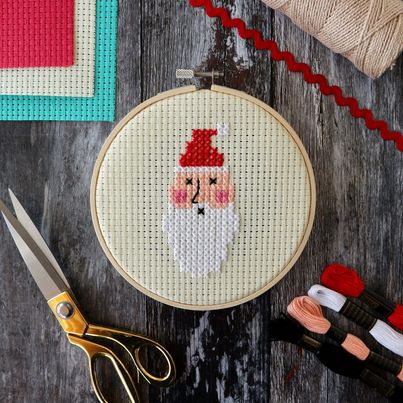Cross Stitch Santa in embroidery hoop on wooden background with string, ribbon, cross Stitch gauze, scissors and threads in foreground.