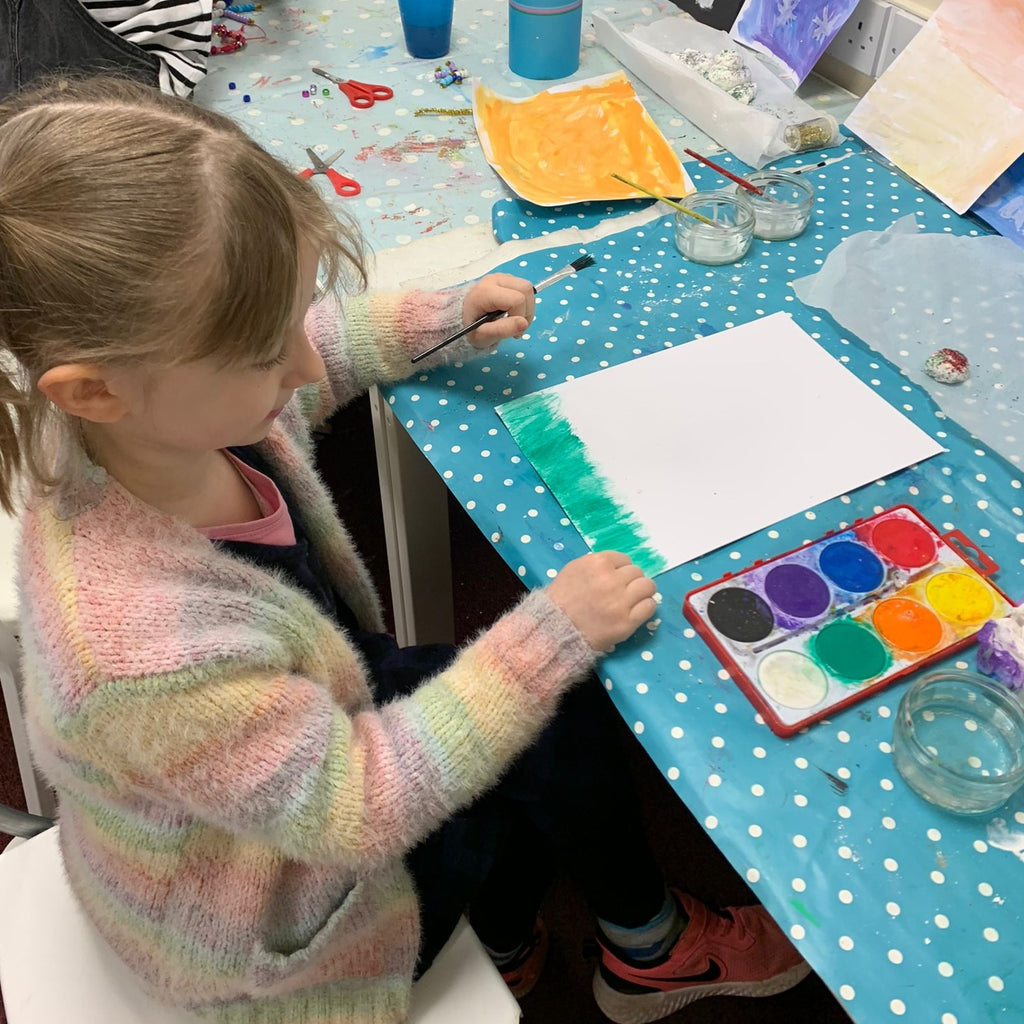 little girl painting using water colour paints, water and paintbrush. Blue polka dot table cloth with craft mess in background.