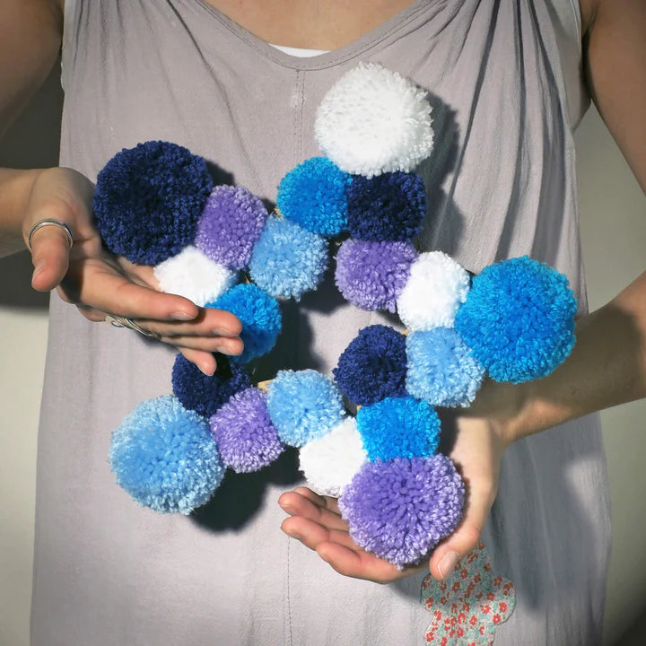 pom pom star wreath in hands. lady holding in her arms wearing mauve jumpsuit and thumb ring.