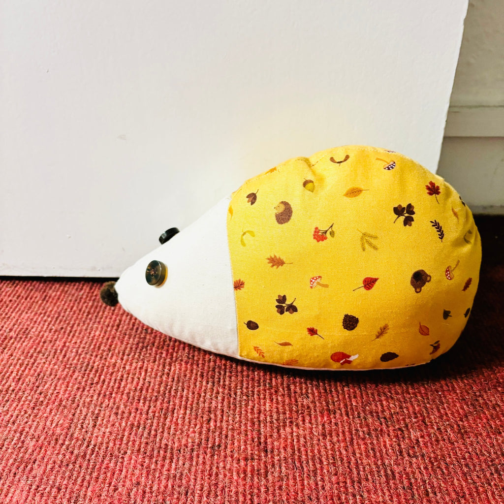 a handmade hedgehog doorstop on red carpet next to white door. button eyes and pom pom nose