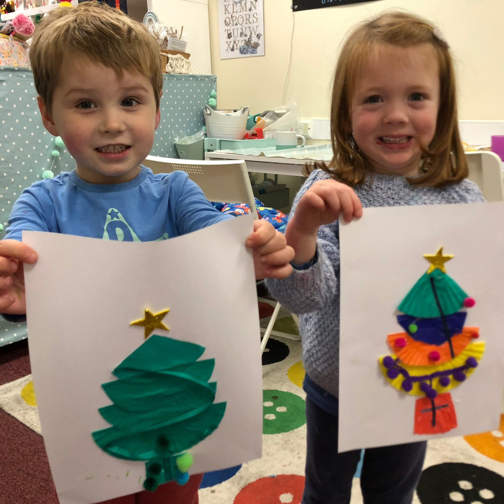little boy and little girl holding up their christmas tree creations on white paper. Button rug and shop in background.