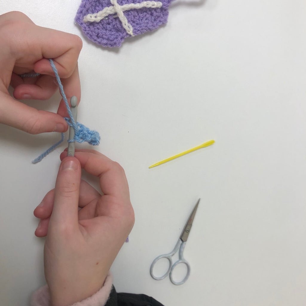 Close up of hands learning to crochet with crochet square and scissors in background.