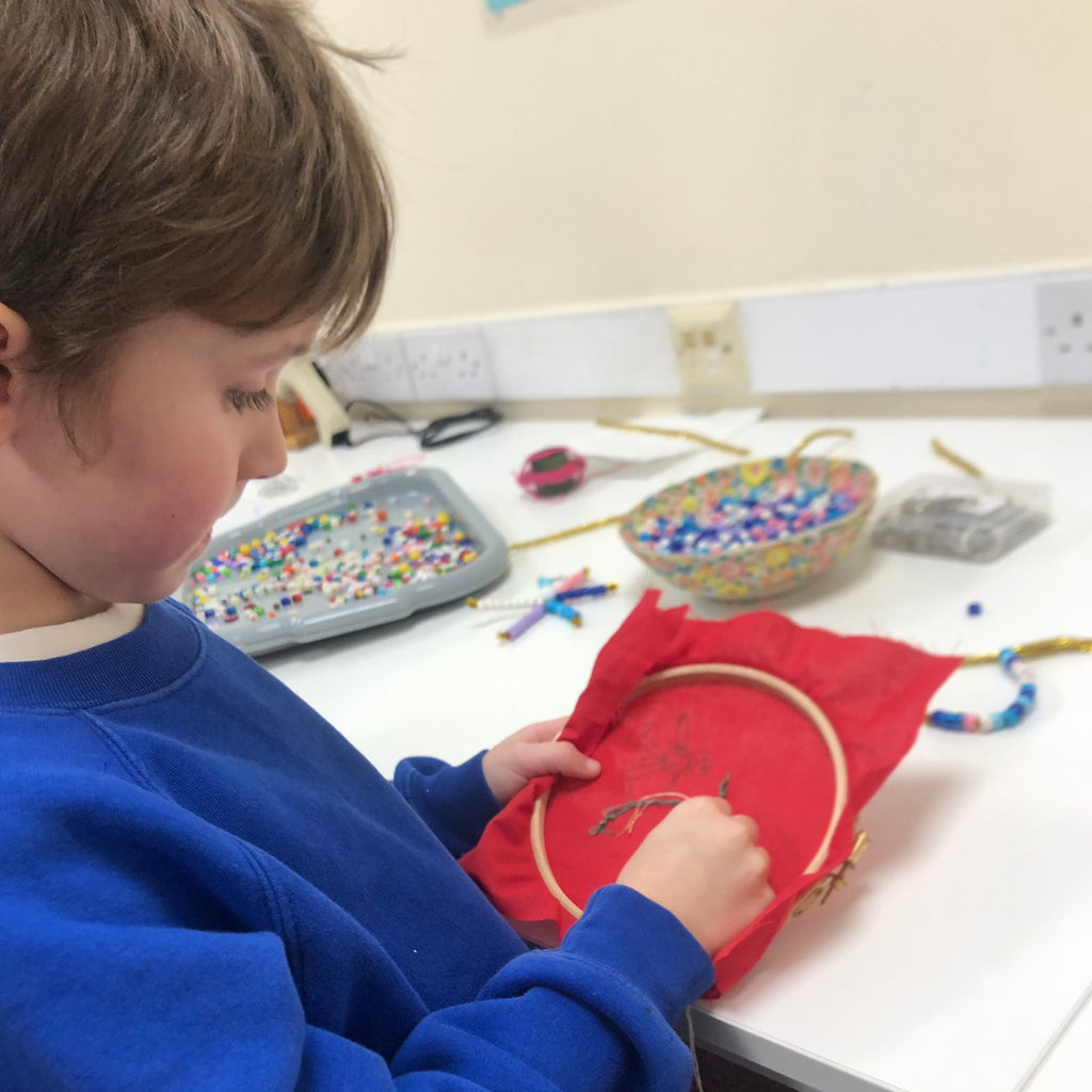 little boy in blue school uniform. He was doing cross stitch with embroidery hoop and red fabric. Beads and bowls of beads in background.