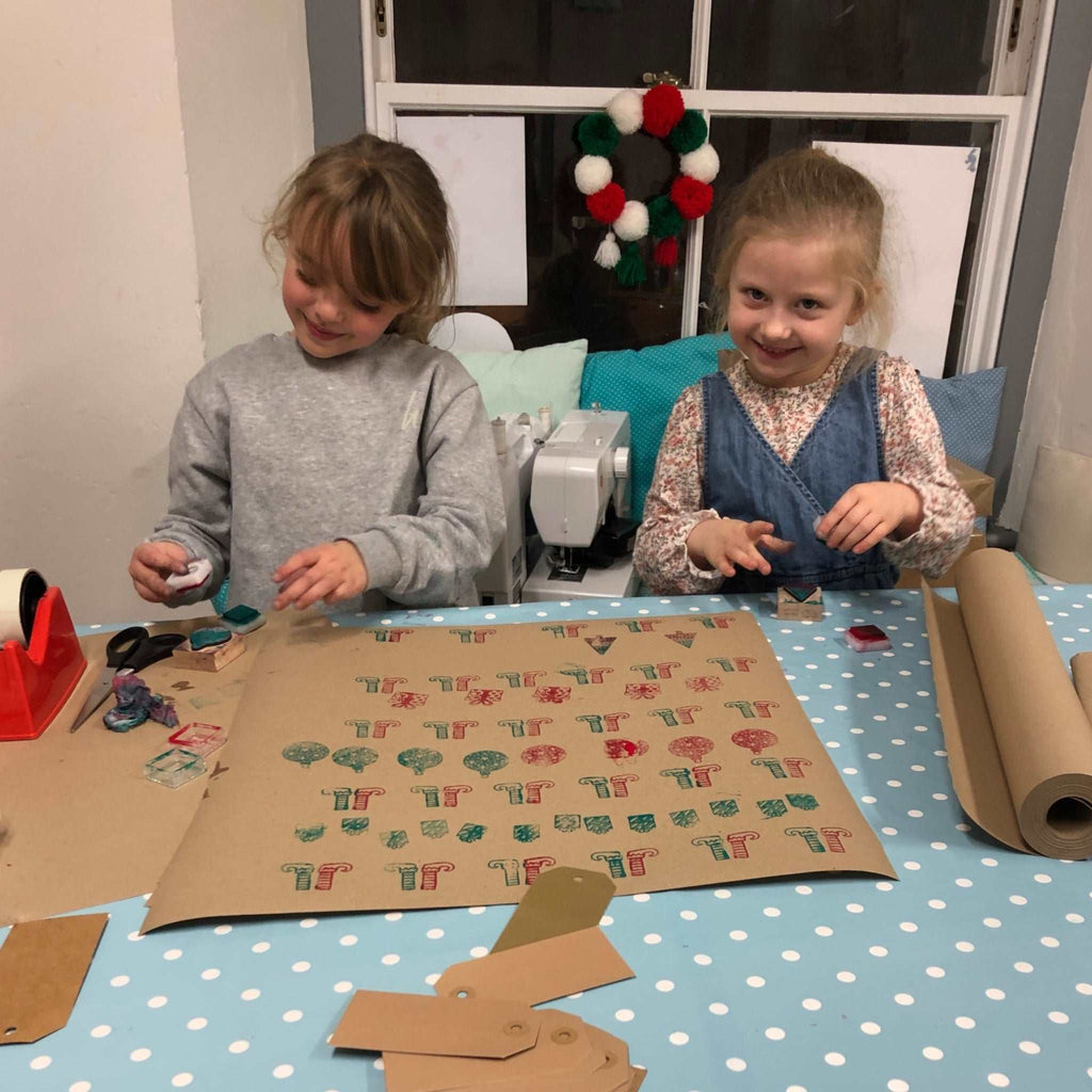 Two primary school aged girls printing Christmas wrapping paper. Brown paper with green and red ink and Christmas stamps.