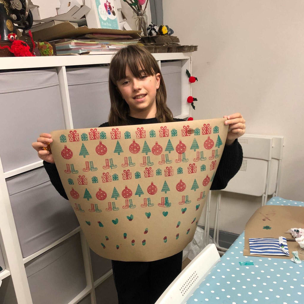 Little girl with dark hair and a fringe holding up her brown printed wrapping paper to show her creation.