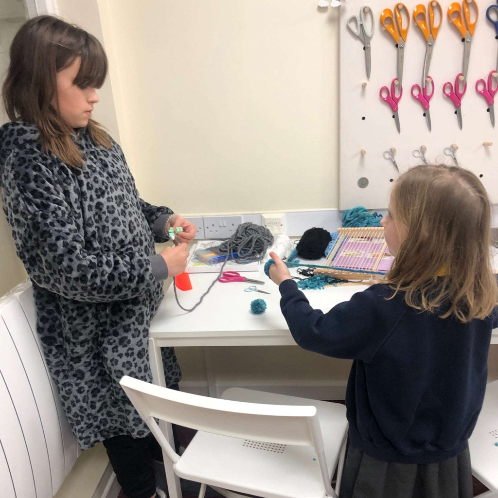 Two young girls making pom poms in craft club. Pegboard of scissors, white table and chairs, weaving loom and wool in background.