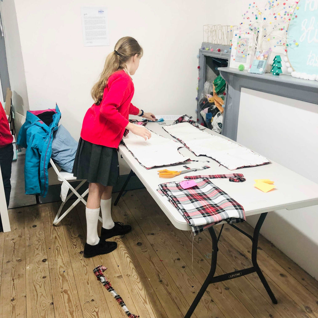 Home Education Stitch Club - little girl in school uniform pattern cutting her tartan outfit on a trestle table.
