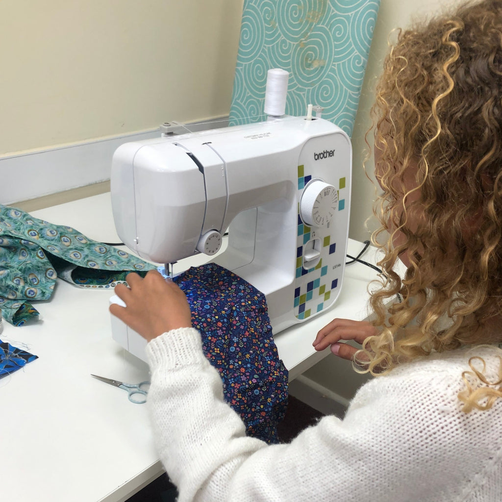 young girl using a sewing machine at stitch club. Blonde curly haired girl using a brother sewing machine to sew blue floral fabric.