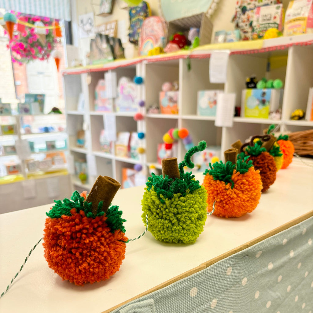 Pom pom pumpkin garland on shop shelf with out of focus craft kit stock in the background.