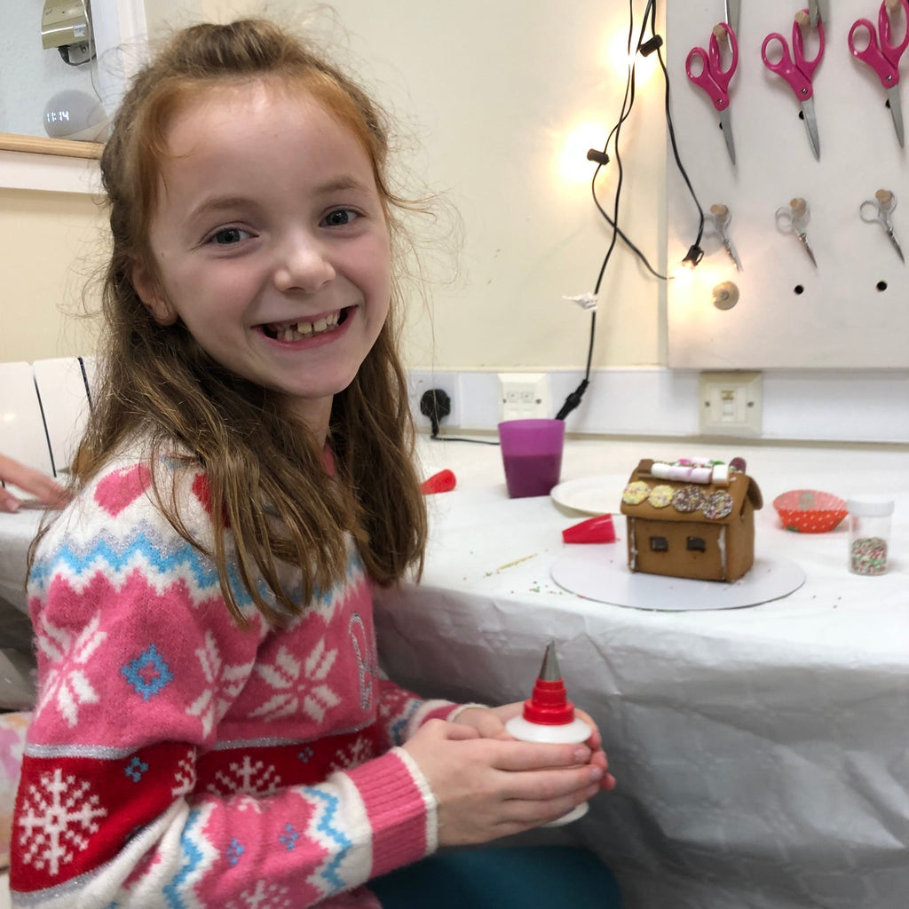 little girl in woollen snowflake christmas jumper with icing pen in hand and half decorated gingerbread house