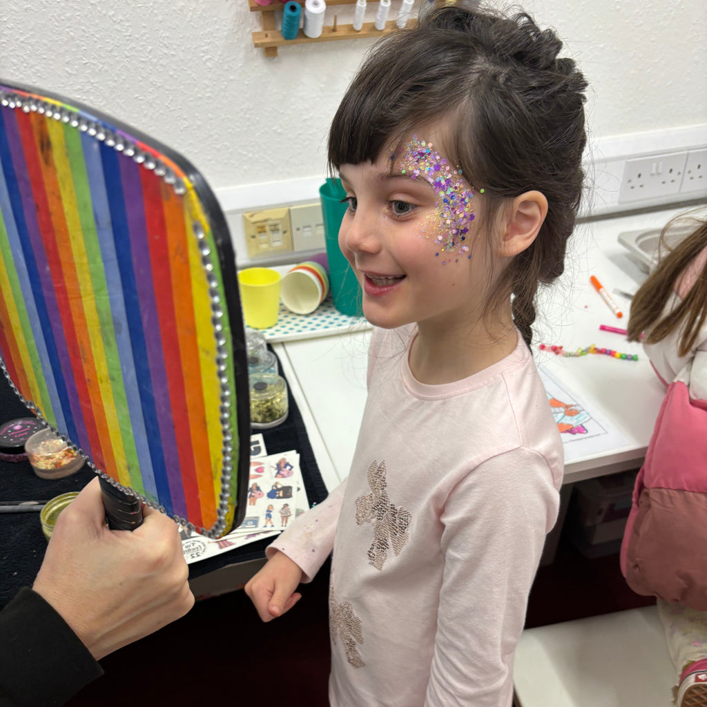 little girl with glitter tattoo on side of face and a mirror held up so she can see