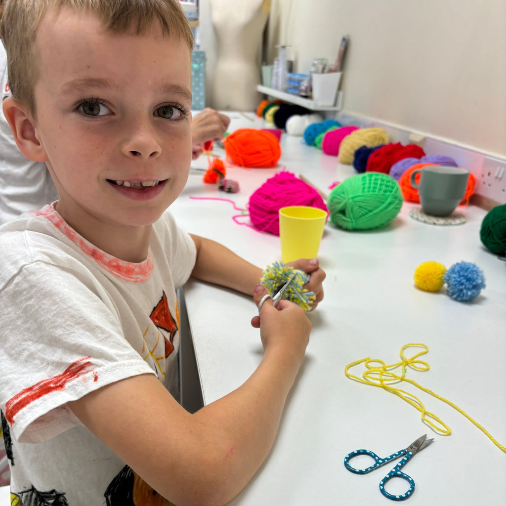 little boy making pom poms with coloured yarn in background.