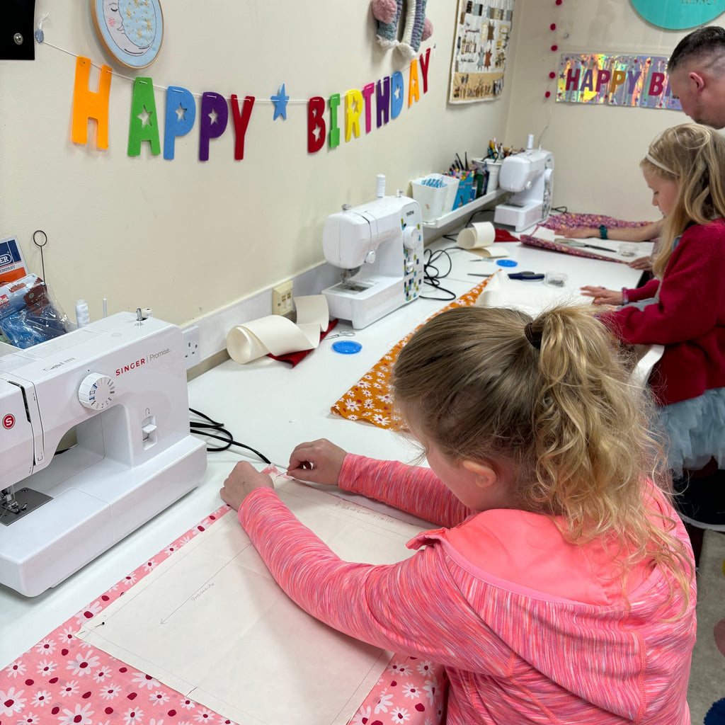 two young girls sewing on sewing machines and tacking their pattern to their floral fabric. happy birthday banner in background.