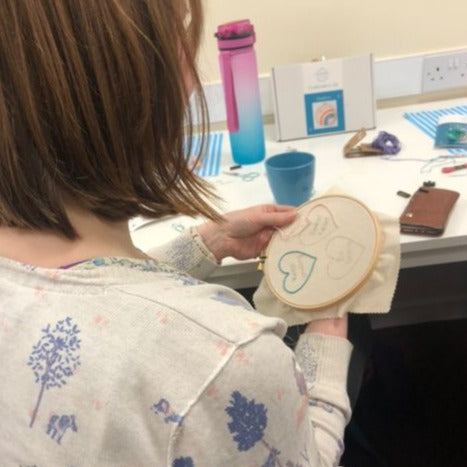 behind view of a lady embroidering hearts in an embroidery workshop with desk in background.